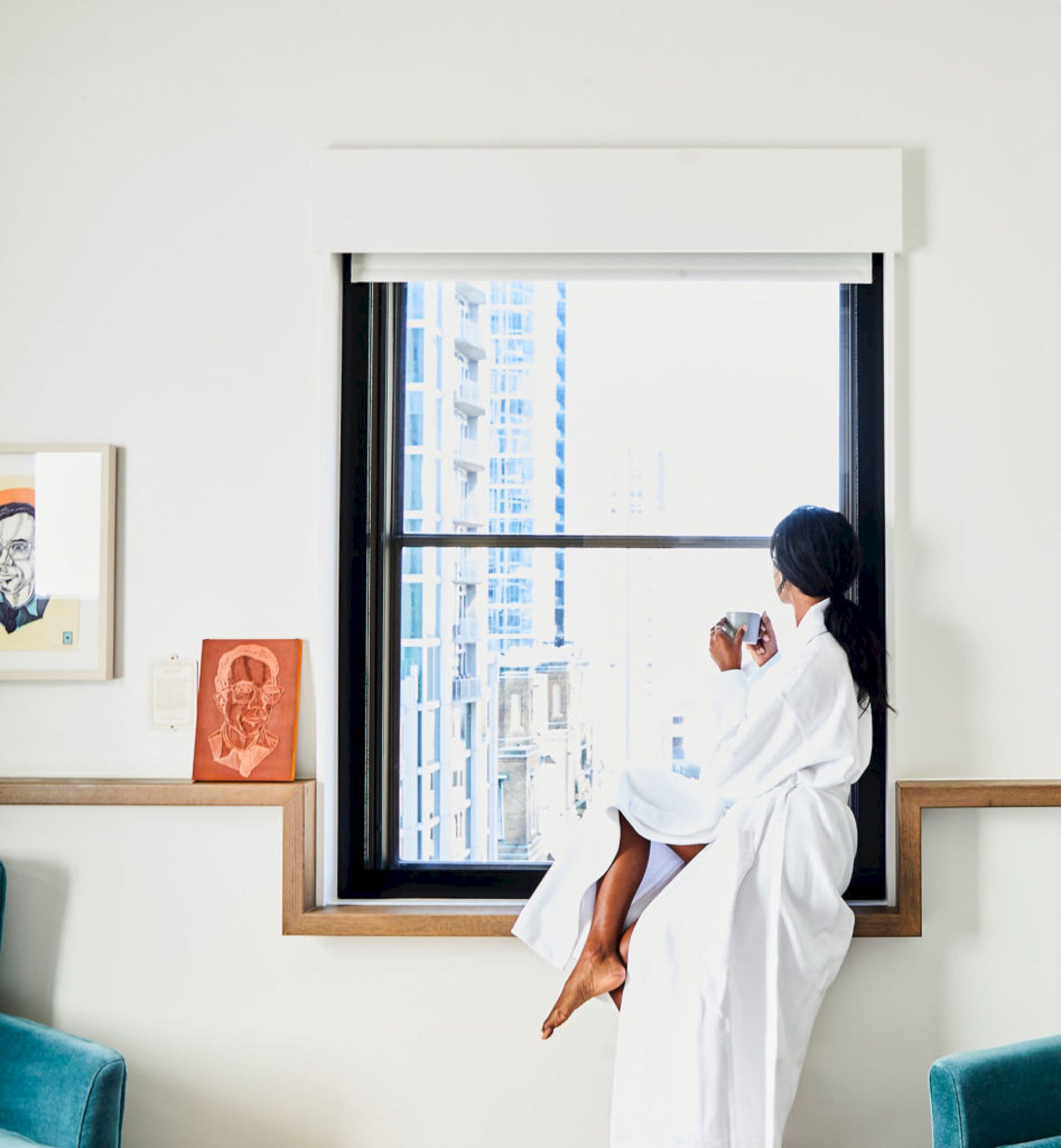 A person in a white robe sits by a window, drinking from a cup, with city buildings visible outside, in a cozy, modern room.