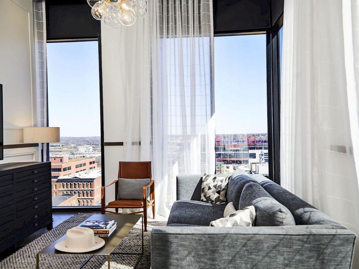 A modern living room with a gray sofa, coffee table, armchair, large windows, sheer curtains, and city view in the background.