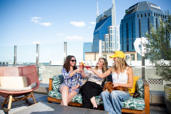 Three people sitting on a rooftop, toasting drinks, with a city skyline in the background.