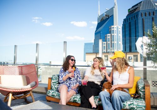 Three people sitting on a rooftop terrace enjoying drinks, with city skyscrapers in the background, featuring a sunny, clear sky.