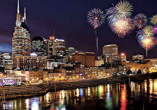 A vibrant city skyline is illuminated at night with colorful fireworks bursting above, reflecting beautifully in the water below.