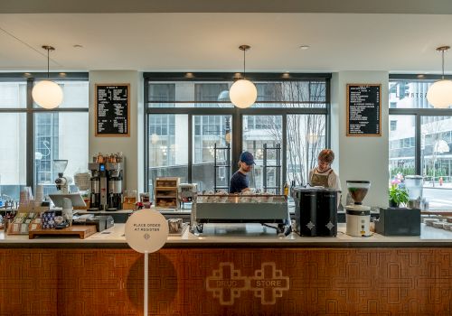 A modern café with two employees behind the counter, coffee equipment, and menu boards on the wall. Natural light fills the space.