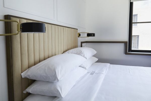 The image shows a neatly made bed with white pillows and sheets, a padded headboard, and wall-mounted lamps in a bright room.