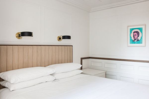 A minimalistic bedroom with a bed, white bedding, wall sconces, and a framed pop art portrait on the wall.