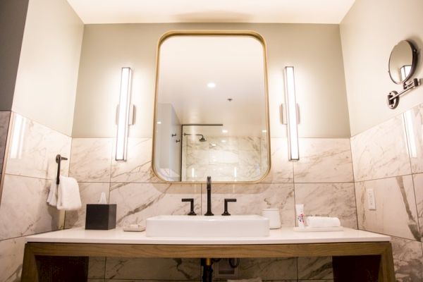A modern bathroom with a large mirror, twin lights, marble walls, a rectangular sink, and a countertop decorated with amenities.