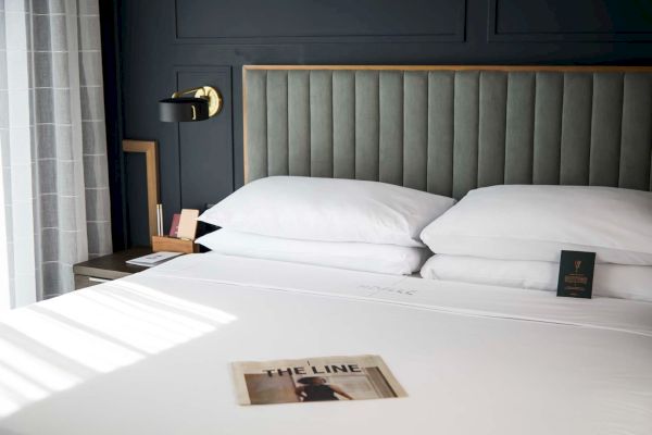 A neatly made bed with four white pillows, a headboard, a side table, and a wall lamp; a magazine titled 
