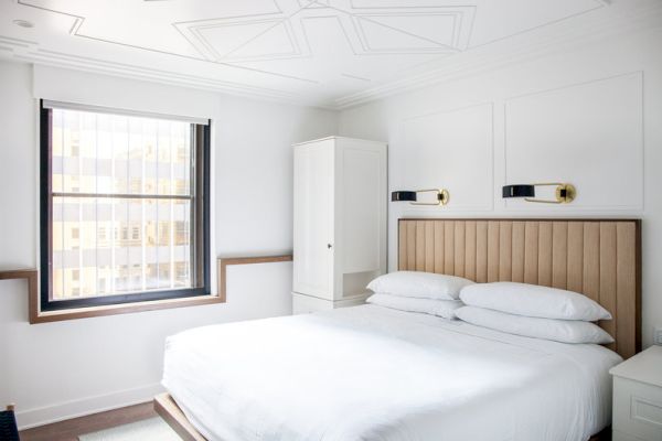 A modern bedroom featuring a neatly made bed, wall-mounted lamps, a window, and a wardrobe in a bright, minimalistic setting.