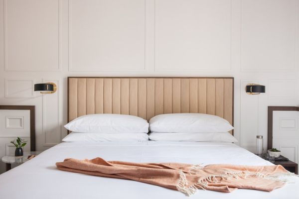 A neatly made bed with white linens and pillows, a tan upholstered headboard, and a peach blanket, flanked by wall-mounted lamps.