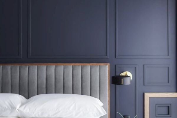 A modern bedroom with a gray upholstered headboard, navy paneled wall, white bedding, and a nightstand with a lamp and small plant.