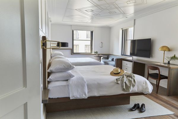 The image shows a hotel room with two beds, a desk, a TV, and a window, featuring a hat and shoes placed on the bed and floor.