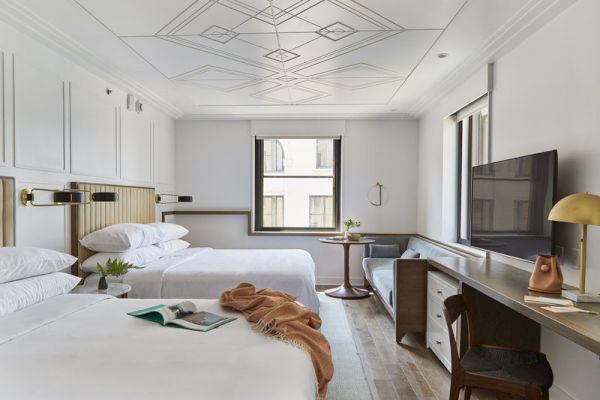 The image shows a modern hotel room with two beds, a TV, a desk, and a large window.