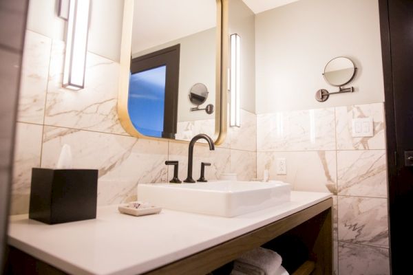 A modern bathroom with a marble countertop, rectangular sink, large mirror, wall-mounted lights, and a tissue box on the counter.
