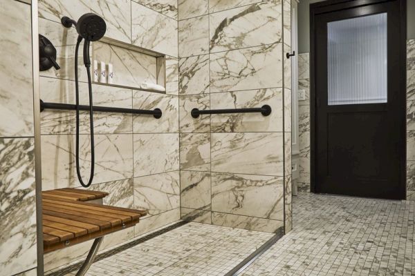 The image shows a marble-tiled shower with a bench, grab bars, a handheld showerhead, and a door leading to a separate area.