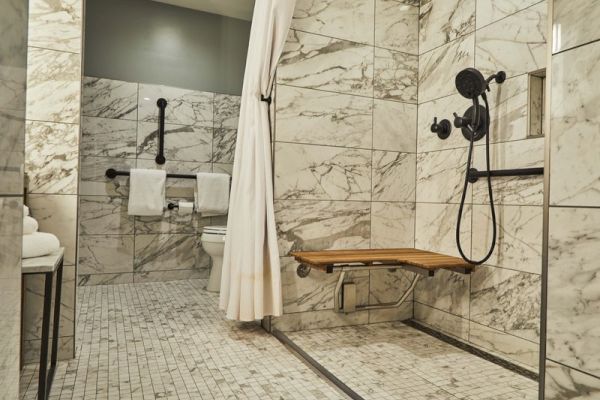 The image shows a marble-tiled accessible bathroom with a shower, a foldable seat, and grab bars for support near the toilet.
