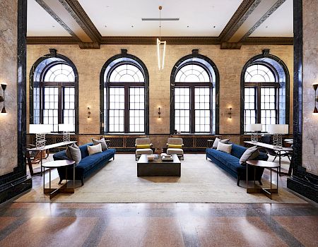 A spacious, elegant room with large arched windows, blue sofas, armchairs, and a coffee table, featuring minimalist decor and soft lighting.