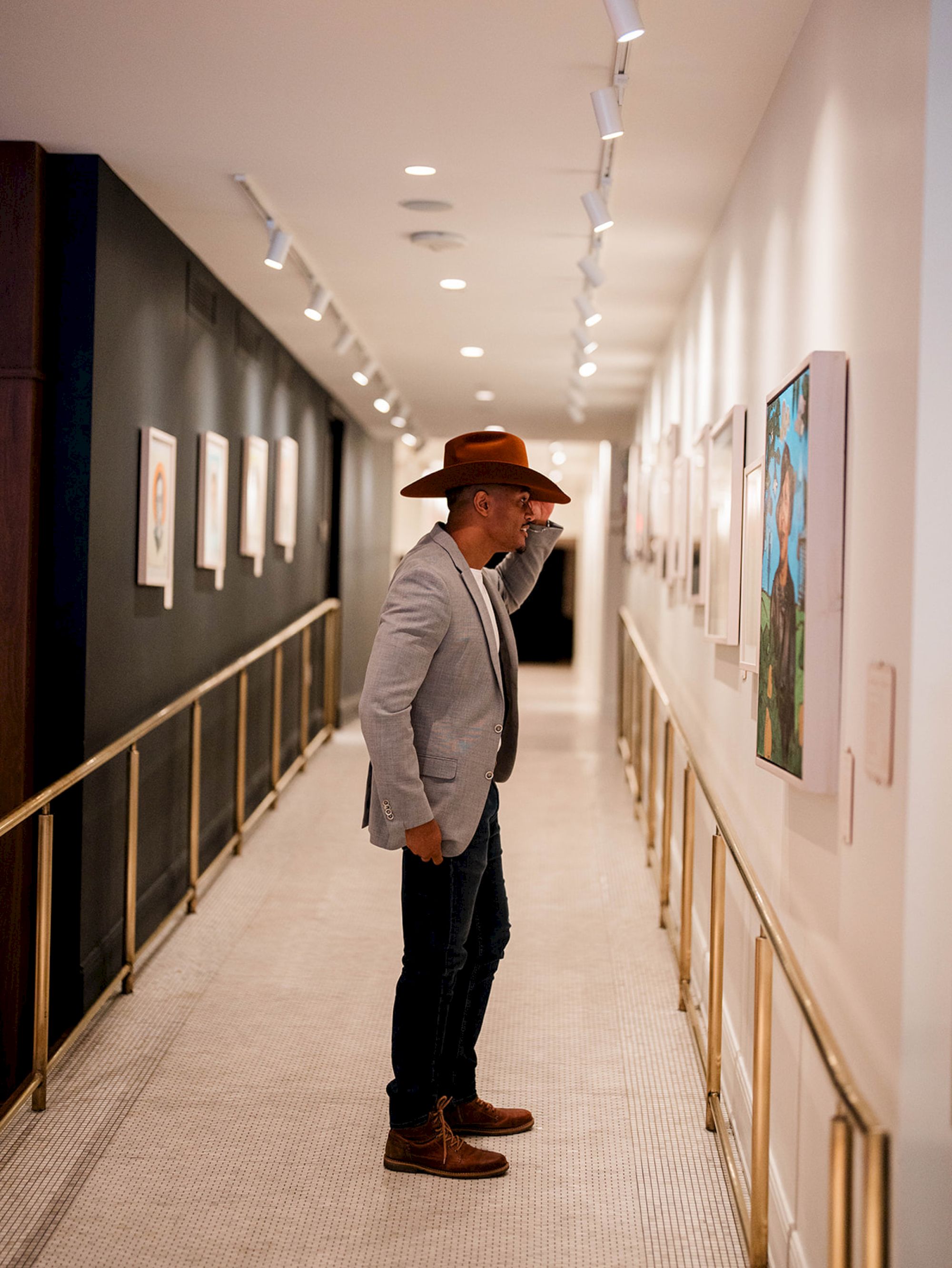 A person wearing a hat and jacket is viewing artwork in a brightly lit hallway with framed pieces on the walls.
