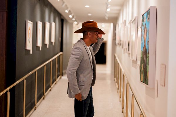 A person in a hat and suit is viewing artwork in a brightly lit gallery hallway, with paintings lined along the walls.