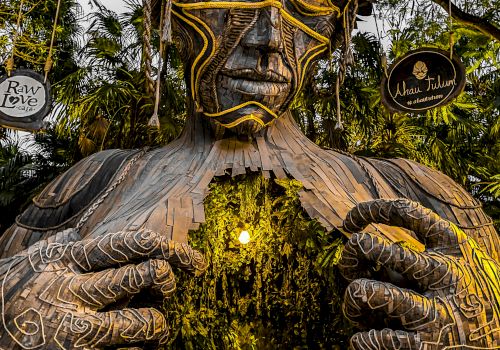 A large, artistic sculpture of a human figure, with intricate patterns, forms an entrance surrounded by greenery in a forest setting.