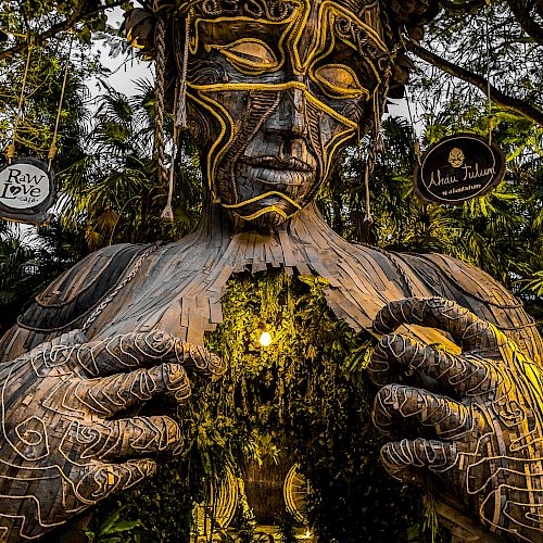 A large, artistic sculpture of a human figure, with intricate patterns, forms an entrance surrounded by greenery in a forest setting.