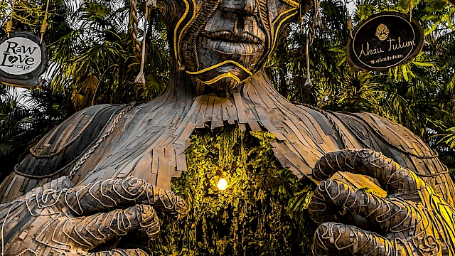 A large, artistic sculpture of a human figure, with intricate patterns, forms an entrance surrounded by greenery in a forest setting.