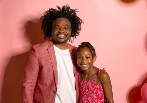 A man and a young girl are smiling in front of a pink backdrop with colorful balloons hanging above.