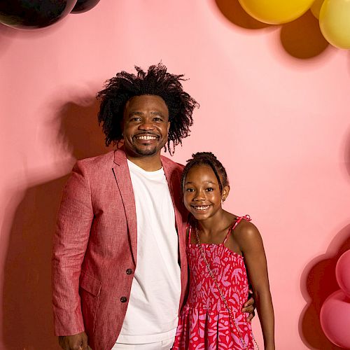 A man and a young girl are smiling in front of a pink backdrop with colorful balloons hanging above.