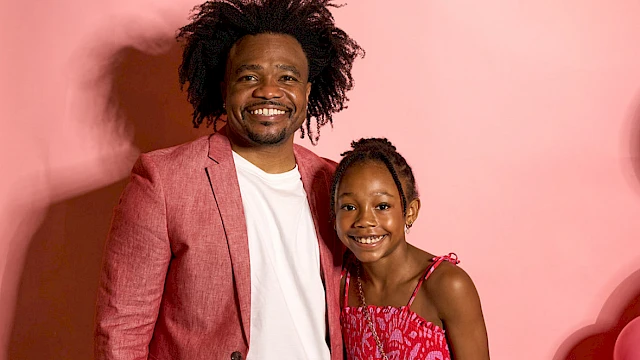 A man and a young girl are smiling in front of a pink backdrop with colorful balloons hanging above.