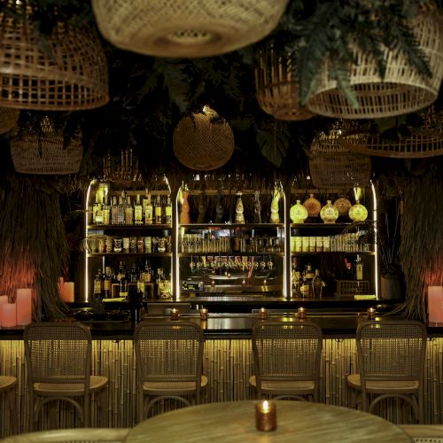 A dimly lit bar with wicker chairs, pendant lighting, and bottles displayed, creating a cozy, inviting atmosphere.