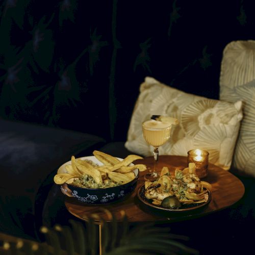 A cozy setting with fries, a cocktail, and a plated dish on a wooden table, beside a candle and cushions.