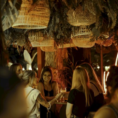 A group of people socialize in a warmly lit, rustic environment with hanging basket-style decor overhead.