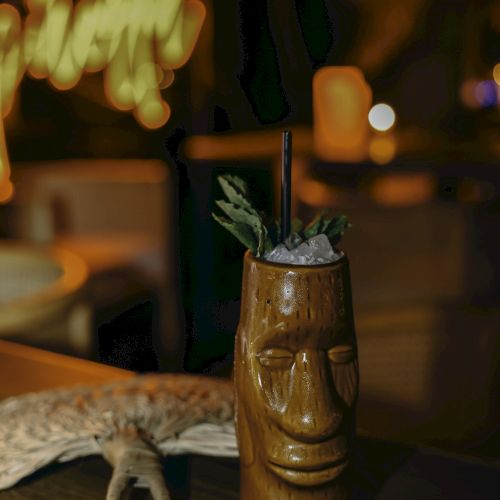 A tiki-style cocktail sits on a table, adorned with herbs and served in a carved wooden mug, with a cozy, dimly lit background.