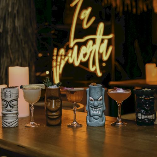 A variety of tiki-style cocktails in colorful mugs lined up on a bar, with candles and a neon 