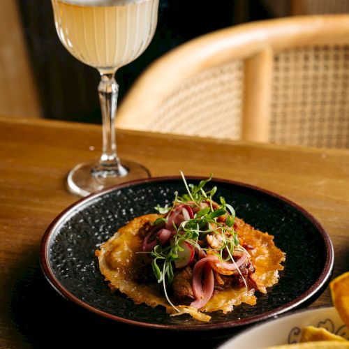A cocktail with a smoking garnish and a plated dish topped with greens on a wooden table next to a chair.