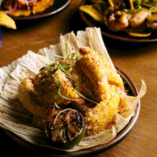 A dish of roasted chicken with garnish sits on a plate, with more food visible in the background on a wooden table.