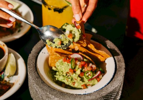 A person is filling tacos with toppings using a spoon. There's a drink nearby and a plate with eggs in the background on the table.