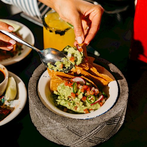 A person is filling tacos with toppings using a spoon. There's a drink nearby and a plate with eggs in the background on the table.