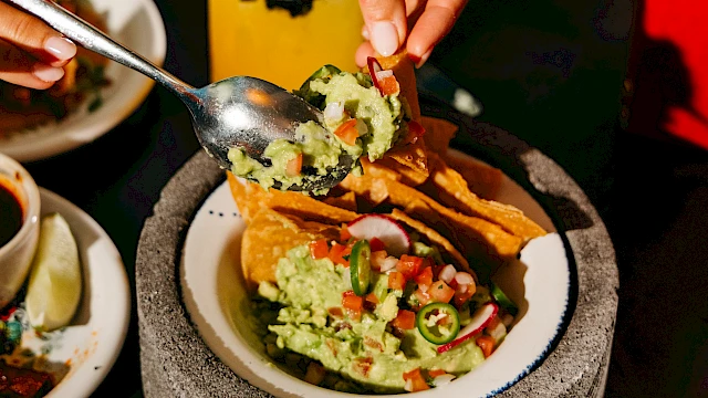 A person is filling tacos with toppings using a spoon. There's a drink nearby and a plate with eggs in the background on the table.