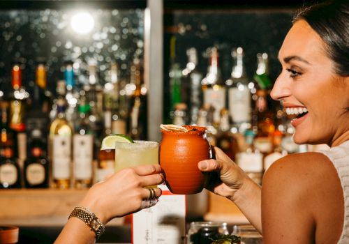 Two people toasting with drinks at a bar, surrounded by bottles and bar items, sharing a joyful moment.