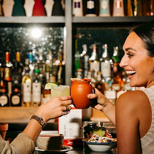 Two people toasting with drinks at a bar, surrounded by bottles and bar items, sharing a joyful moment.