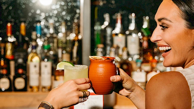 Two people toasting with drinks at a bar, surrounded by bottles and bar items, sharing a joyful moment.