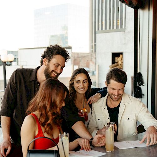 A group of four people gathered around a bar table, looking at menus, with drinks and a handbag on the table.