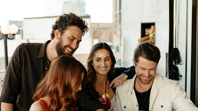 A group of four people gathered around a bar table, looking at menus, with drinks and a handbag on the table.